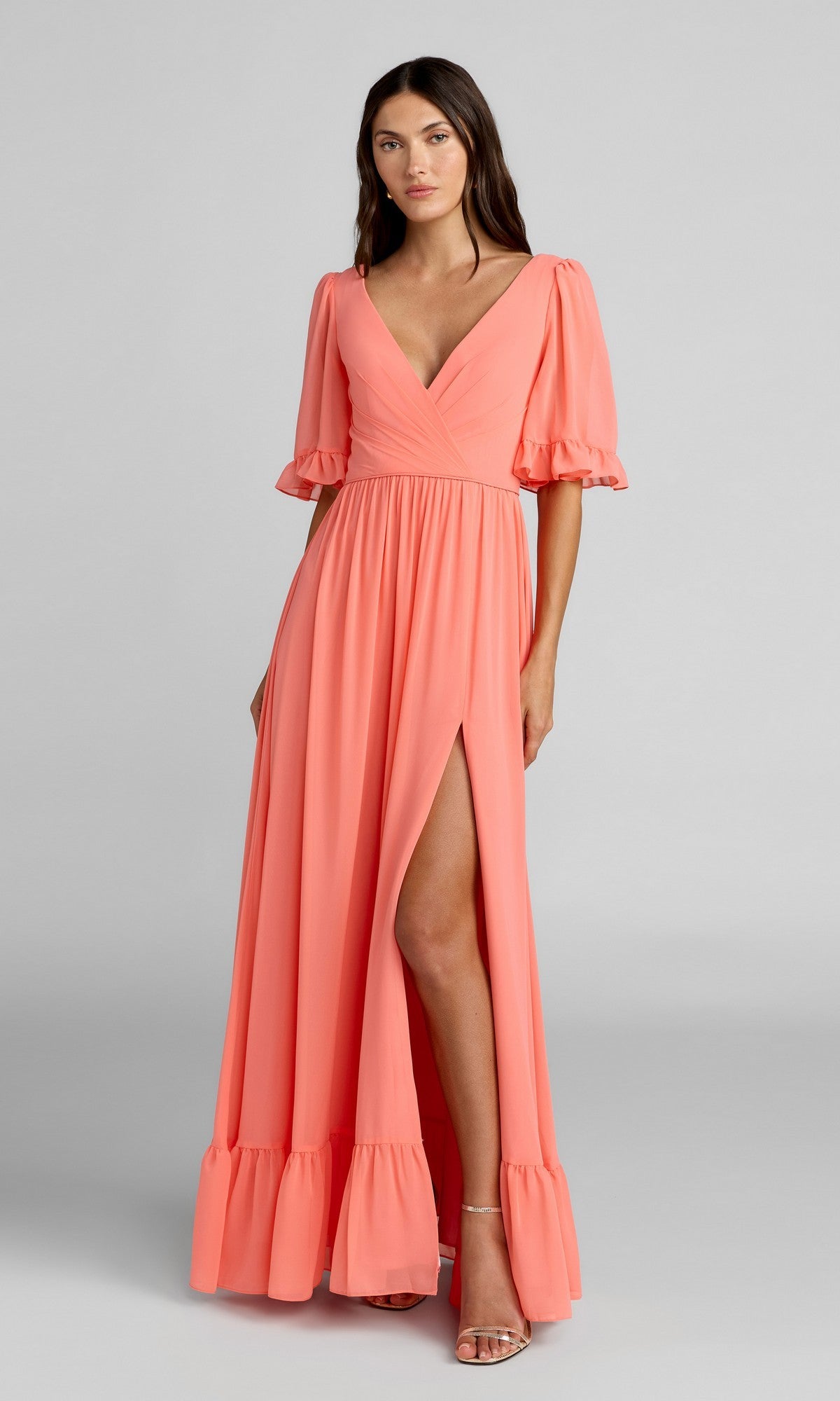 A woman models the Long Formal Dress 55867 by Mac Duggal—a coral pink, floor-length A-line gown with puffed short sleeves, deep V-neckline, front slit, and ruffle hem—while standing in heels against a plain background.